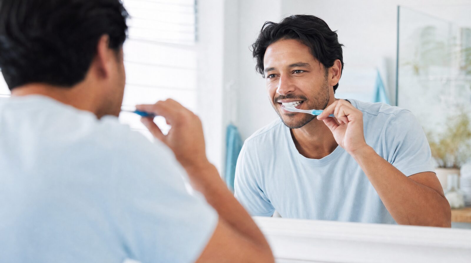 A man brushing his teeth in front of a bright bathroom mirror in soft morning light